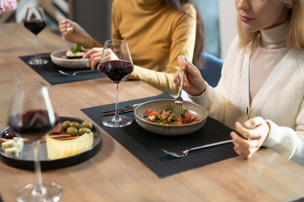 Young blond female in smart casualwear sitting by served table against her friend and having vegetable salad, red wine, cheese and olives