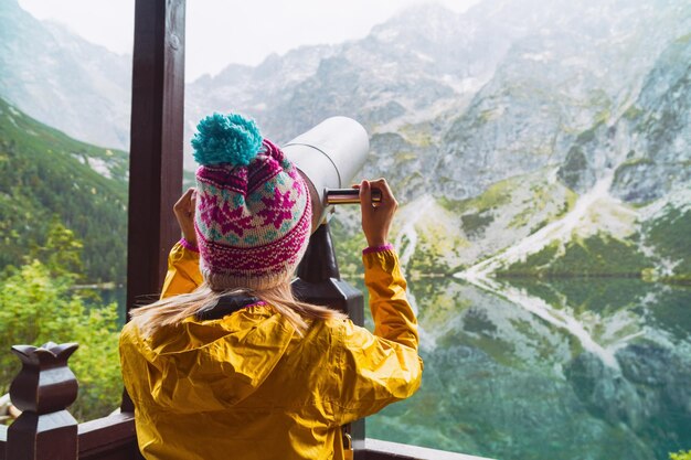 Young blond hair woman wearing yellow jacket purple and blue cap looking through a coin operated binoculars on mountains and lake Copy space rear view background