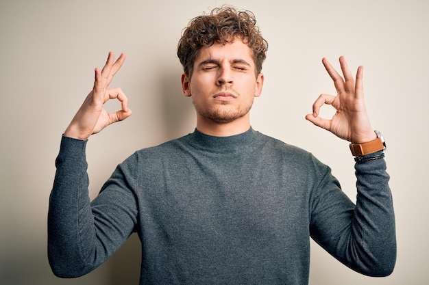 Young blond handsome man with curly hair wearing casual sweater over white background relax and smiling with eyes closed doing meditation gesture with fingers Yoga concept