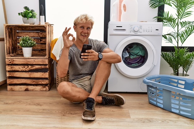 Young blond man doing laundry using smartphone smiling positive doing ok sign with hand and fingers successful expression