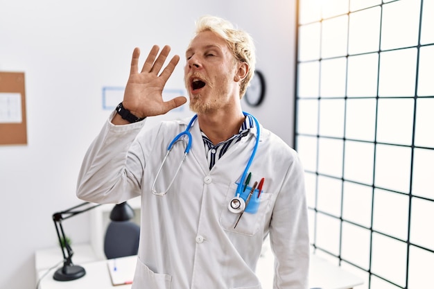 Young blond man wearing doctor uniform and stethoscope at clinic shouting and screaming loud to side with hand on mouth. communication concept.