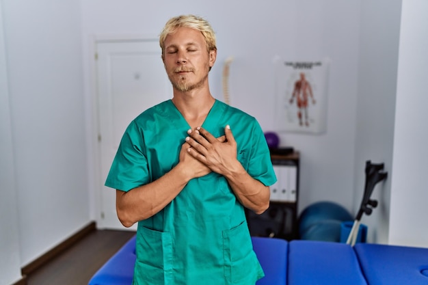 Young blond man wearing physiotherapist uniform standing at clinic smiling with hands on chest with closed eyes and grateful gesture on face. health concept.