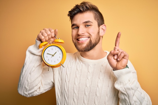 Young blond man with beard and blue eyes holding alarm clock over yellow background surprised with an idea or question pointing finger with happy face number one