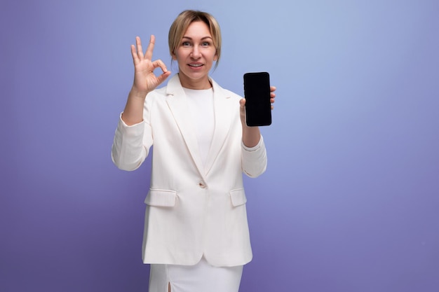 Young blond manager woman in a white elegant jacket and skirt shows the screen of a smartphone with