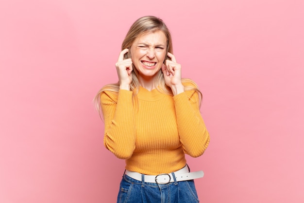 Young blond pretty woman looking angry, stressed and annoyed, covering both ears to a deafening noise, sound or loud music