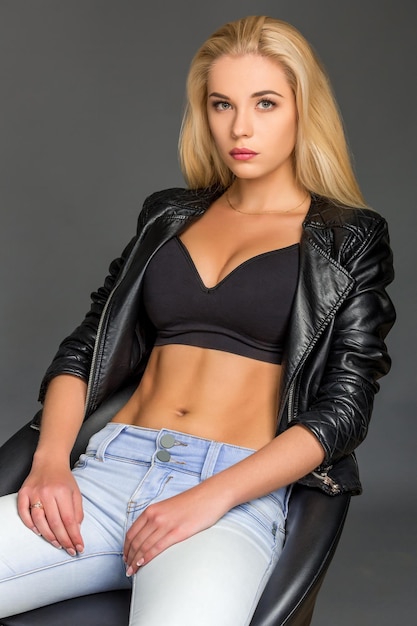 young blond woman in a black leather jacket, blue jeans and a top. she is sitting on a chair and looking at the camera