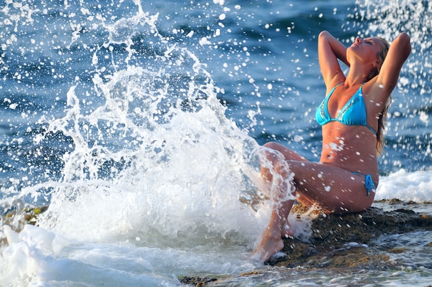 Young blond woman in blue bikini sitting on rocks and enjoying drops of waves on clear sunny summer day. Happiness, vacations and freedom concept