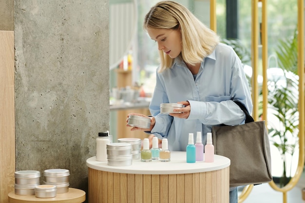 Young blond woman choosing body cream or scrub in tin jar in supermarket