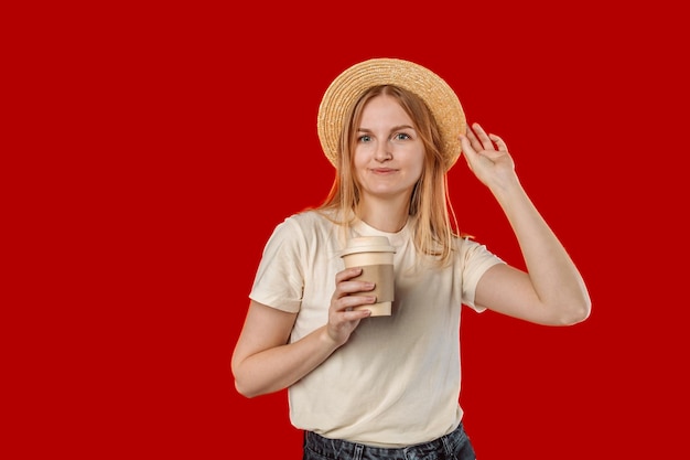 Young blond woman in hat with a paper eco coffee to go over on red color background studio portrait