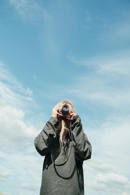 Young blond woman in khaki shirt with a hood holding camera in front of blue sky outdoors