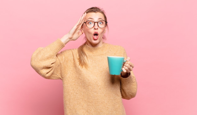 Young blond woman looking happy, astonished and surprised, smiling and realizing amazing and incredible good news. coffee concept