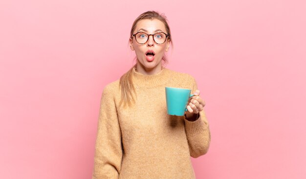 Photo young blond woman looking very shocked or surprised, staring with open mouth saying wow. coffee concept