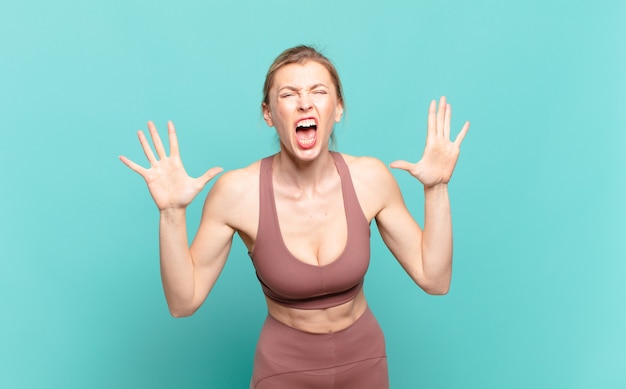 Young blond woman screaming in panic or anger, shocked, terrified or furious, with hands next to head. sport concept