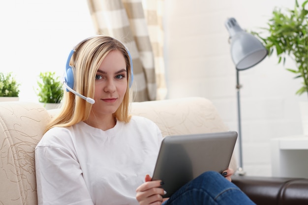 young blond woman sit on the sofa in livingroom hold tablet in arms listen to music sunny