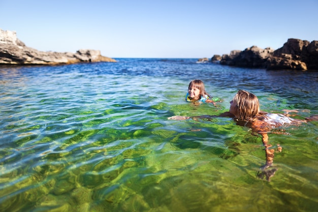 Young blond woman and small happy girl swimming and having fun in clear sea water with rocks at background on clear summer day