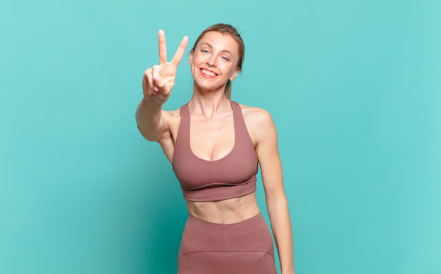 Young blond woman smiling and looking happy, carefree and positive, gesturing victory or peace with one hand. sport concept