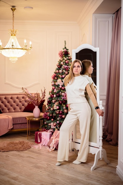 Young blond woman in a white knitted suit standing in front of a mirror in front of a Christmas tree Winter holidays new year