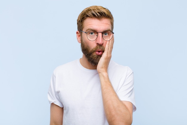 Young blonde adult man feeling shocked and astonished holding face to hand in disbelief with mouth wide open