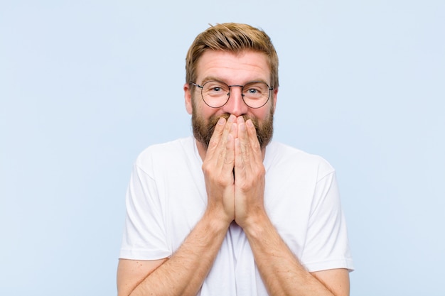 Young blonde adult man happy and excited, surprised and amazed covering mouth with hands, giggling with a cute expression