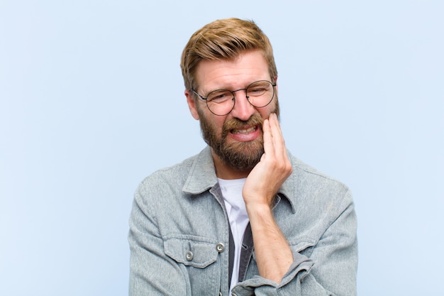 Young blonde adult man holding cheek and suffering painful toothache, feeling ill, miserable and unhappy, looking for a dentist