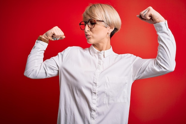 Young blonde business woman with short hair wearing glasses over red background showing arms muscles smiling proud Fitness concept