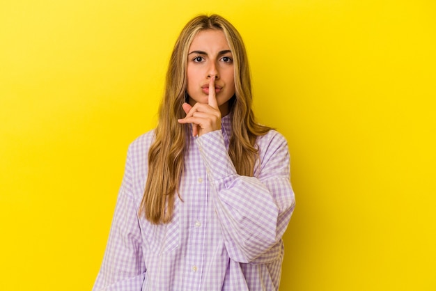 Young blonde caucasian woman isolated on yellow wall keeping a secret or asking for silence.