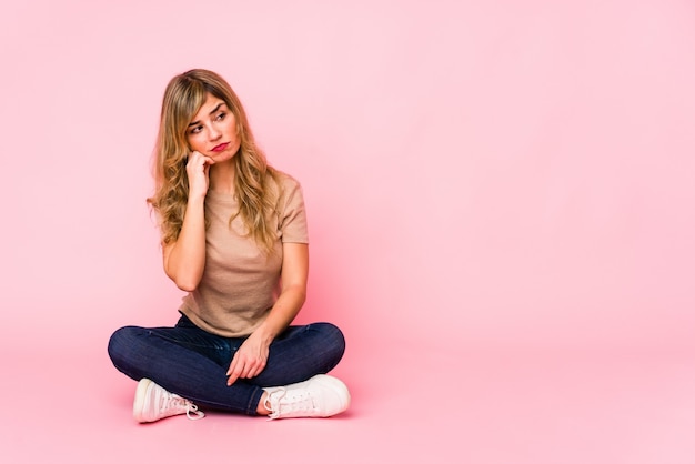 Photo young blonde caucasian woman sitting on a pink studio who feels sad and pensive, looking at blank space.