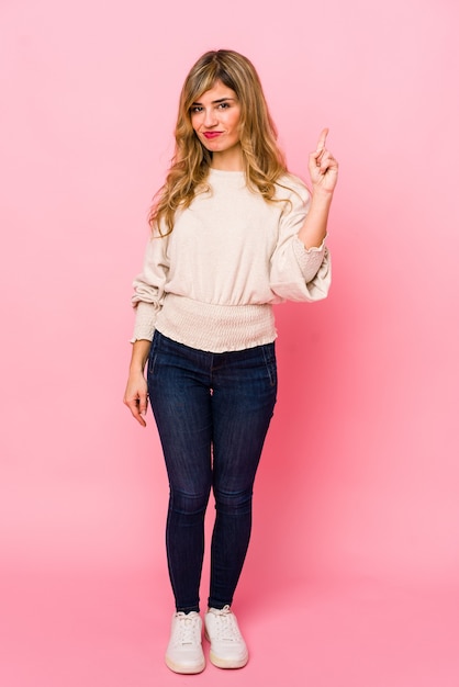 Young blonde caucasian woman standing over a pink background showing number one with finger.