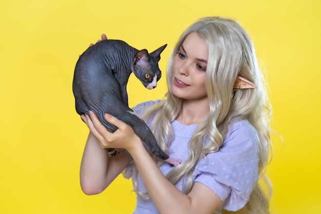 Young blonde cosplay elf in blue dress with curly hair holding Sphinx kitten in hands and smiles