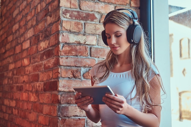 Young blonde female listening music and using a digital tablet while sitting on a window sill in a room with a loft interior.