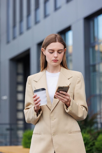 A young blonde girl against the backdrop of a business center office center Portrait of a successful startup Smartphone coffee casual suit