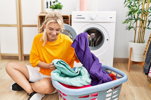 Young blonde girl doing laundry putting clothes into washing machine at home