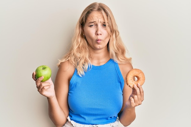 Young blonde girl holding green apple and donut making fish face with mouth and squinting eyes, crazy and comical.