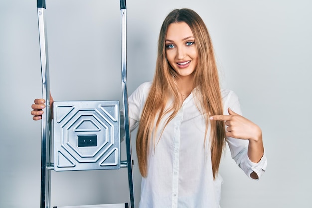 Young blonde girl holding ladder pointing finger to one self smiling happy and proud