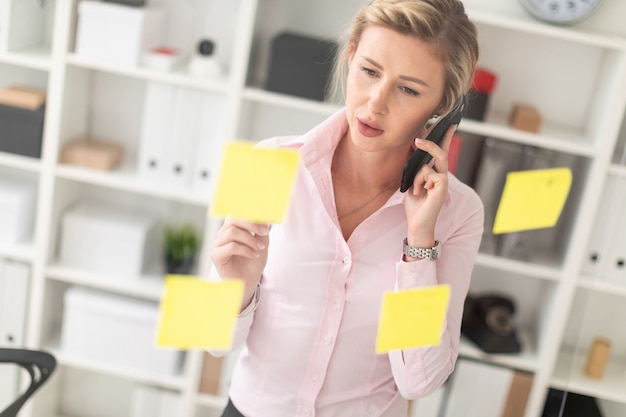 A young blonde girl is standing in the office next to a transparent board with stickers, holding a pencil in her hand and talking on the phone.