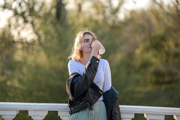 A young blonde girl in a leather jacket drinks coffee in summer