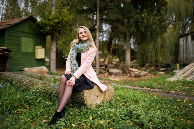 Young blonde girl at pink coat posed on autumn park, sitting on cut tree against house of the forester.