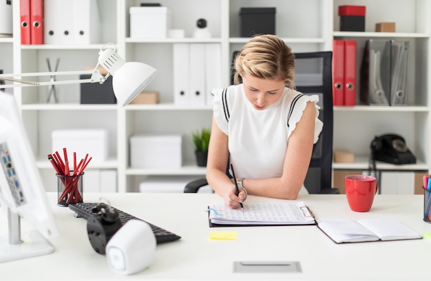 A young blonde girl sits at a computer desk in the office and writes a pencil in the document.