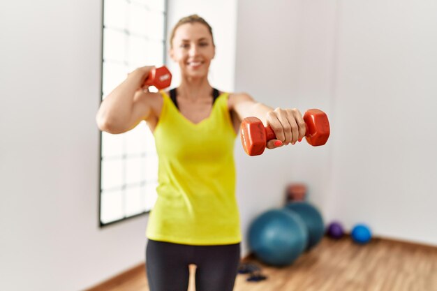 Young blonde girl smiling happy training using dumbbells at sport center.