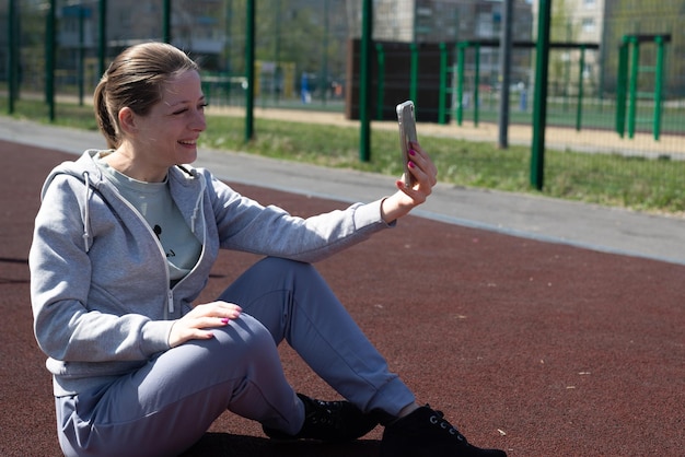 A young blonde girl in sportswear is resting after doing sports running sitting at the stadium talking on a mobile phone social networks