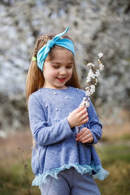 Young blonde girl standing in a blooming garden. Blooming cherry. Portrait of beautiful little girl. Close up of little model face. Sakura blooming, spring evening time