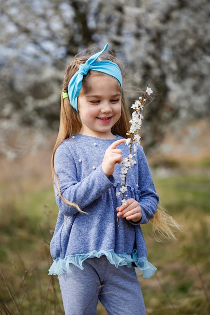 Young blonde girl standing in a blooming garden. Blooming cherry. Portrait of beautiful little girl. Close up of little model face. Sakura blooming, spring evening time