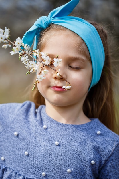 Young blonde girl standing in a blooming garden. Blooming cherry. Portrait of beautiful little girl. Close up of little model face. Sakura blooming, spring evening time