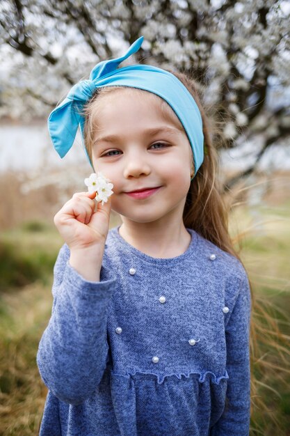 Young blonde girl standing in a blooming garden. Blooming cherry. Portrait of beautiful little girl. Close up of little model face. Sakura blooming, spring evening time