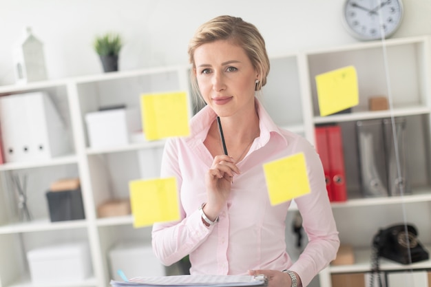 A young blonde girl stands in the office next to a transparent board with stickers and holds documents