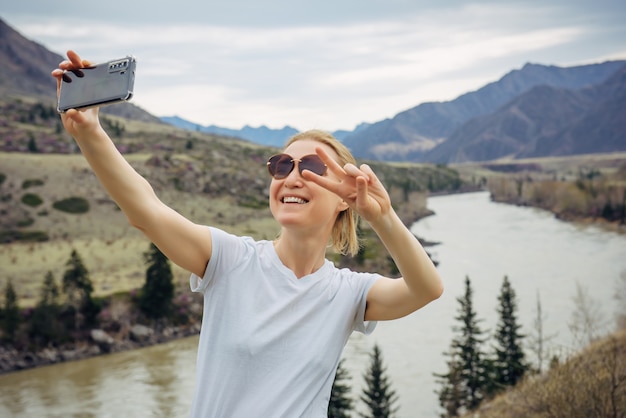 Young blonde girl in sunglasses and white T-shirt takes a selfie on her smartphone on the background of river and rocky mountains. Happy female tourist smiles at mobile phone camera.