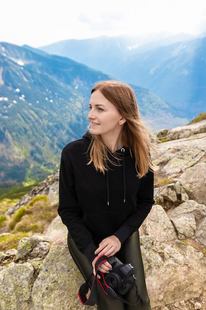 Young blonde girl traveler with a blue backpack raised and enjoying the green mountain scenery 