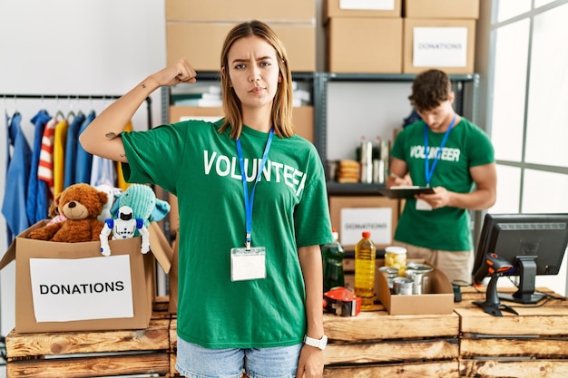 Young blonde girl wearing volunteer t shirt at donation stand strong person showing arm muscle confident and proud of power