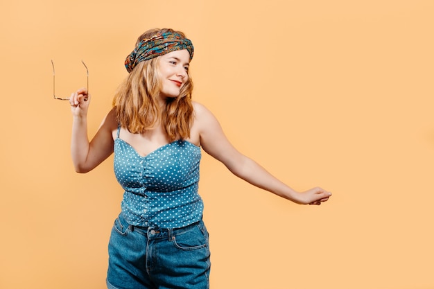 young blonde girl with a bright headscarf dancing holding sunglasses on a light orange background