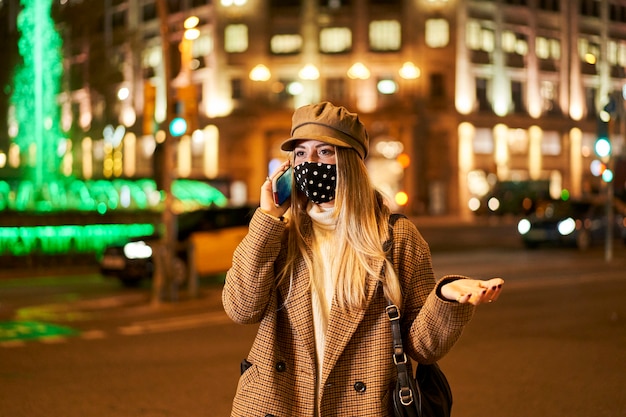 Young blonde girl with mask talking on the phone, gesturing with her hands. She is in a city at night. Winter atmosphere.